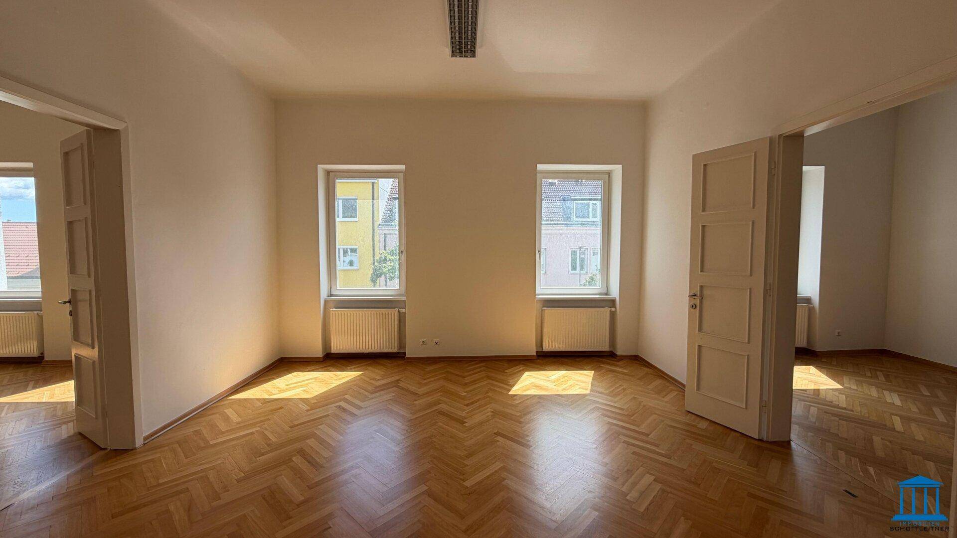 Großes, helles Zimmer mit Fischgrätparkett, zwei Fenstern und Zugang zu weiteren Räumen.