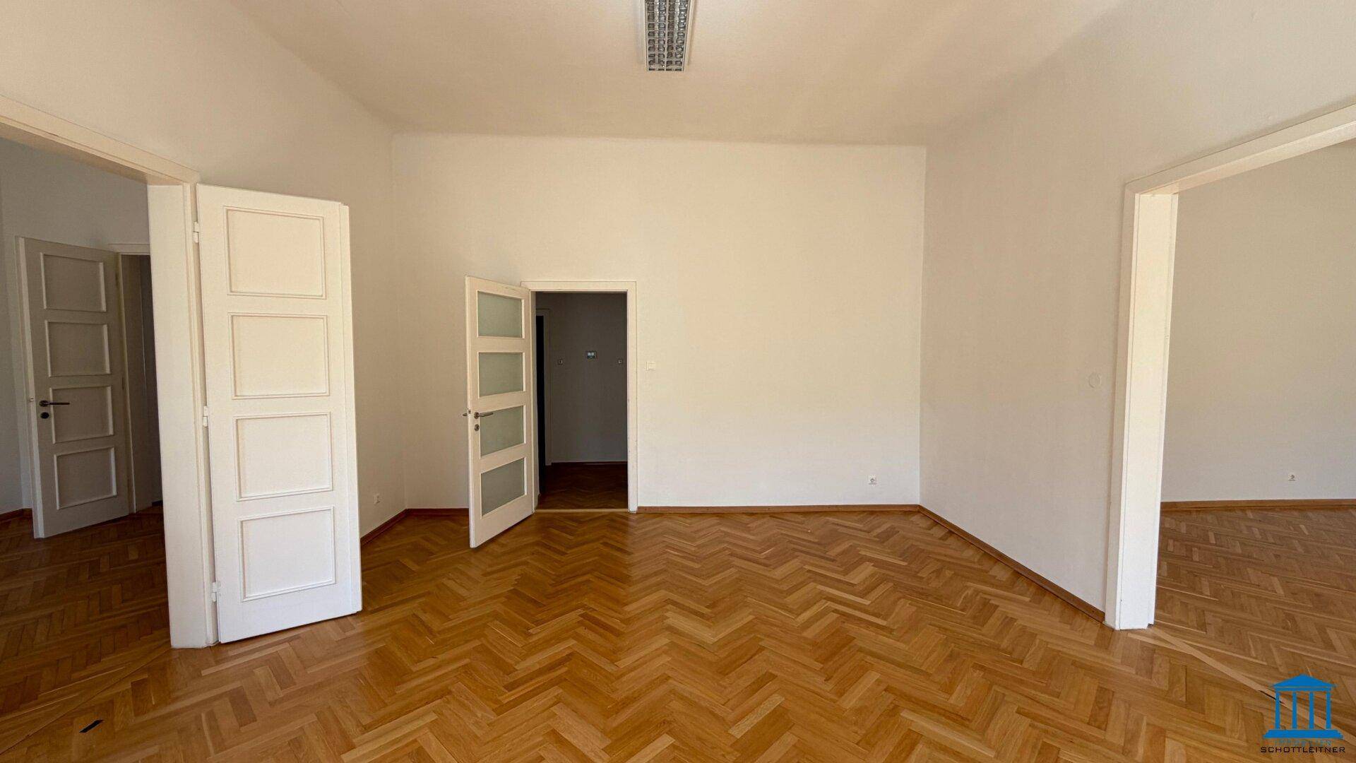 Leeres Zimmer mit Fischgrätparkettboden und mehreren Türen, die zu anderen Bereichen führen.