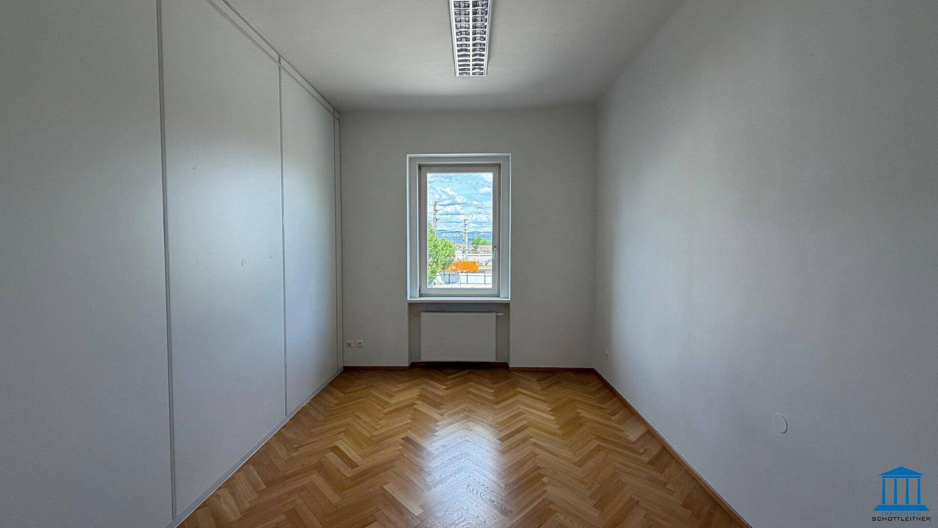 Leeres Büro mit Fischgrätparkett, einem Fenster mit Grünblick und Trennwänden an den Seiten.