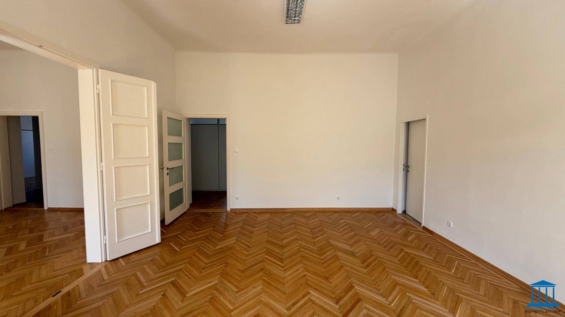 Helles, leeres Zimmer mit Fischgrätparkett und mehreren Türen zu angrenzenden Räumen.