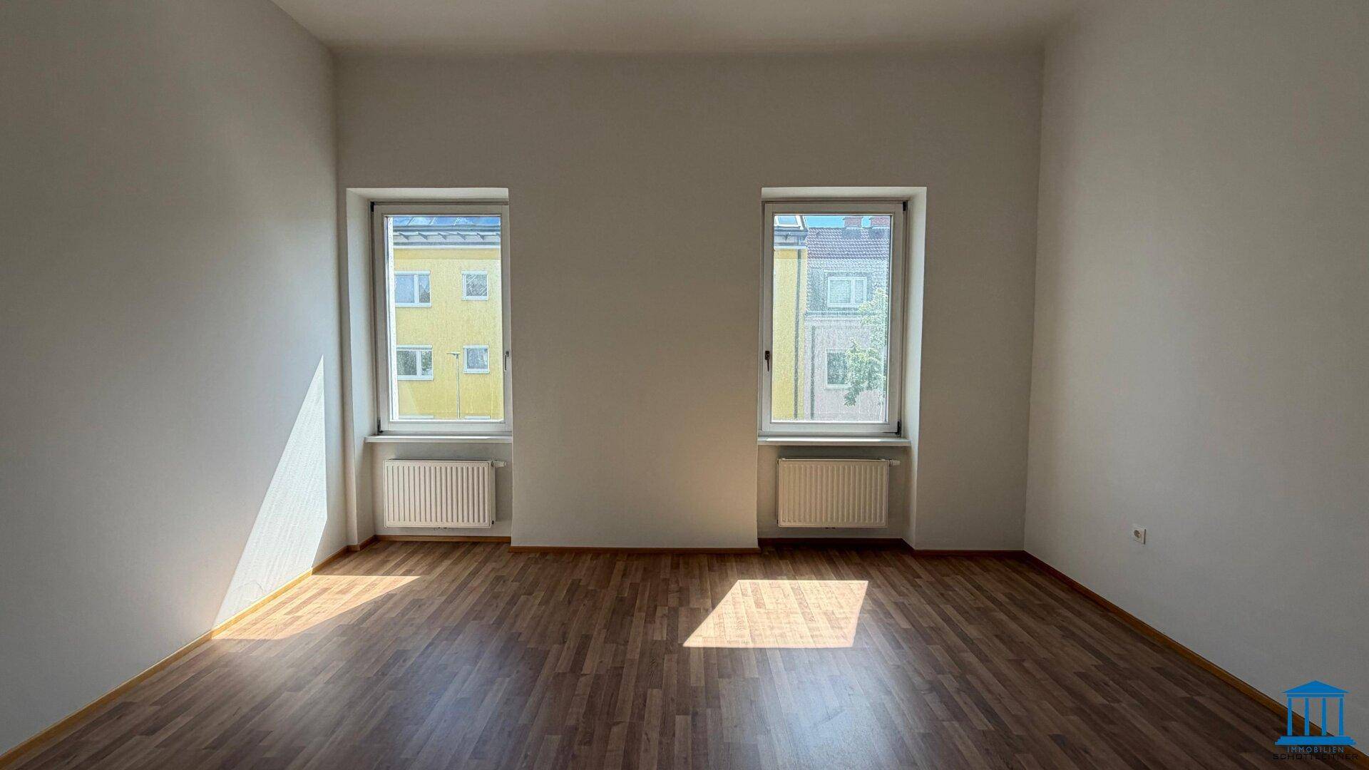 Sonniger Raum mit zwei Fenstern, die den Laminatboden erhellen, und Heizkörpern unter den Fenstern.