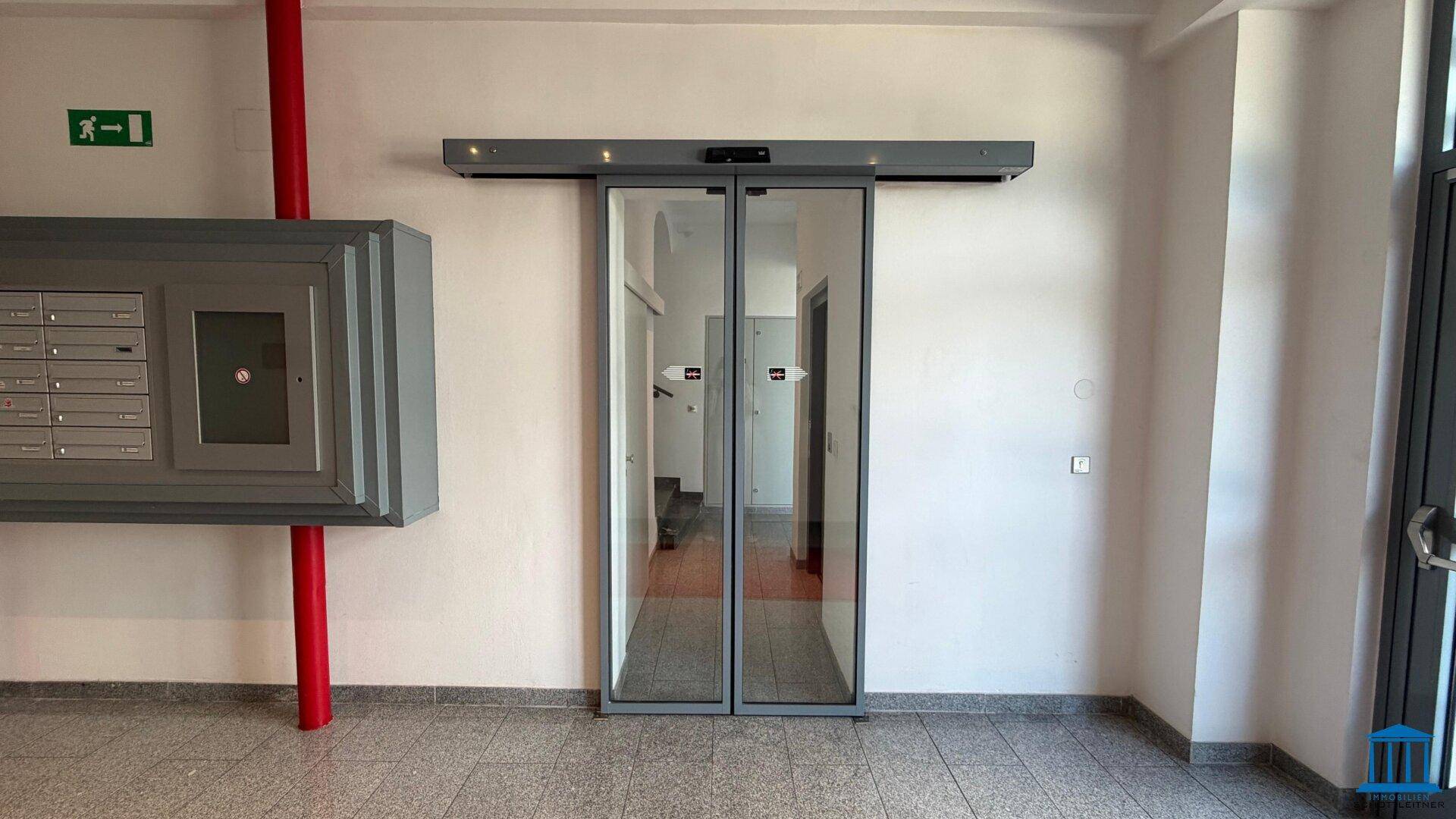 Eingangsbereich mit automatischen Glastüren, Briefkästen und Fliesenboden, hell und einladend.