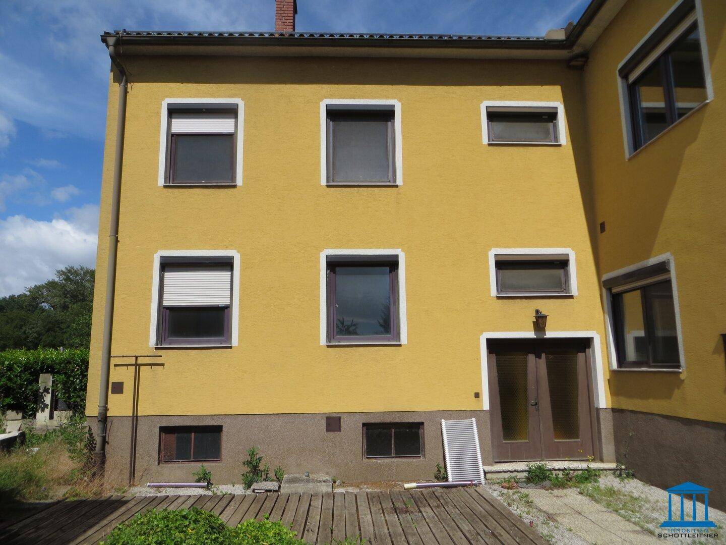 Rückseite des Hauses mit gelber Fassade, Rollläden und einer kleinen Holzterrasse im Erdgeschoss.