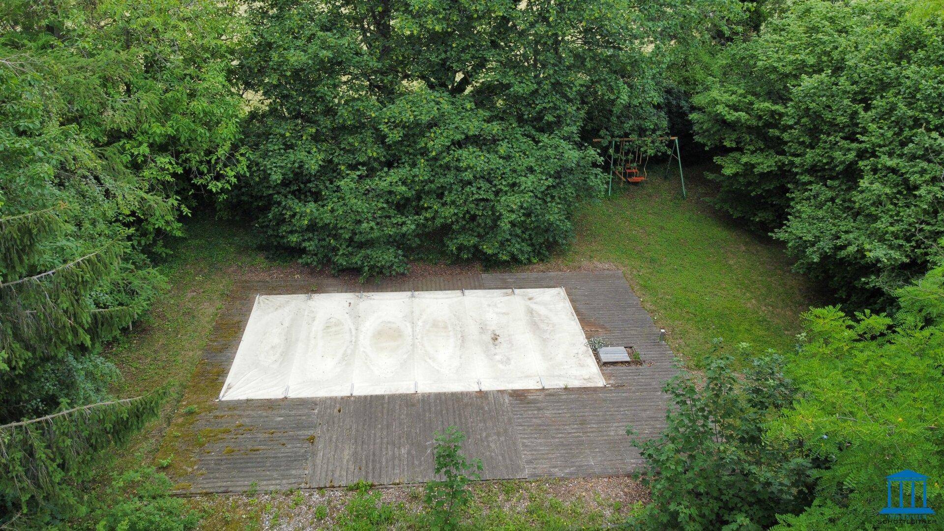 Überdachter Poolbereich im Garten, umgeben von Bäumen und einer Schaukel im Hintergrund.