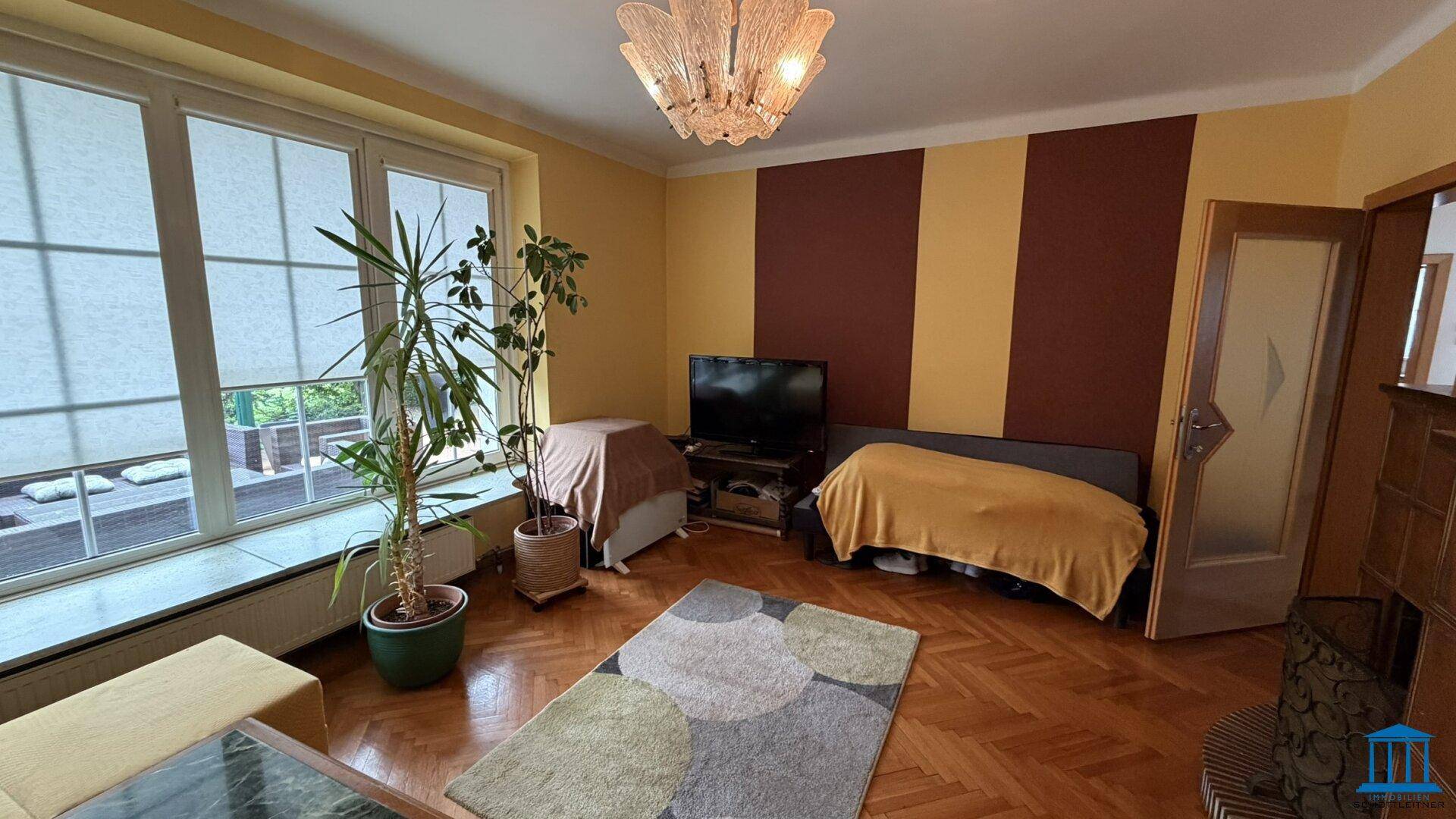 Wohnzimmer mit Fernseher, Parkettboden und großem Fenster, das viel Tageslicht hereinlässt.