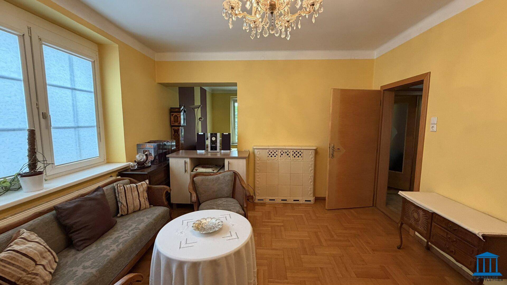 Wohnzimmer mit Parkettboden, Fenster und einem Durchgang zu einem weiteren Bereich.
