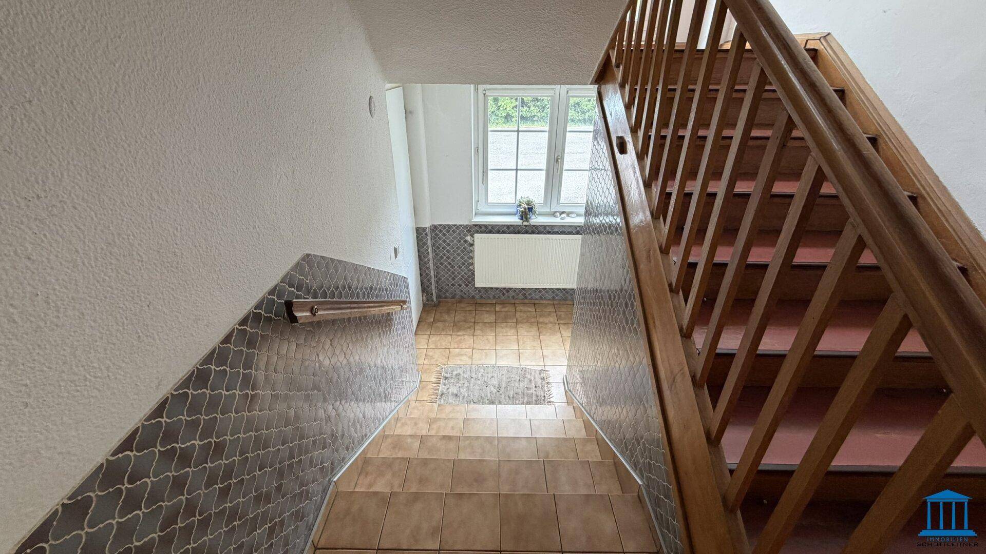 Holztreppe mit Fliesenboden im Treppenhaus, Fenster und Heizkörper im unteren Bereich.