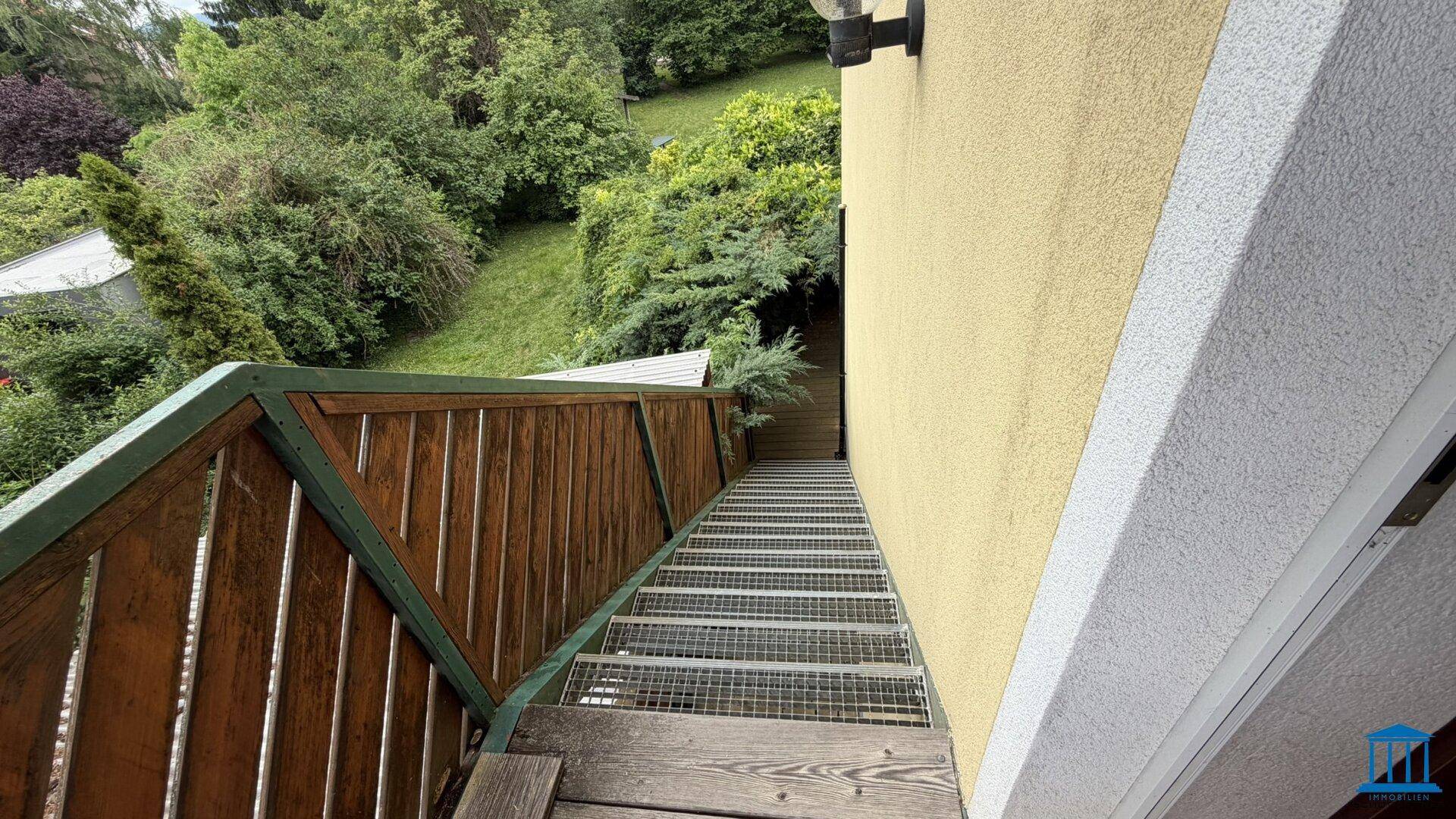 Außentreppe mit Holzgeländer und Metallstufen, die zum Gebäude hinaufführt, umgeben von üppigem Grün.