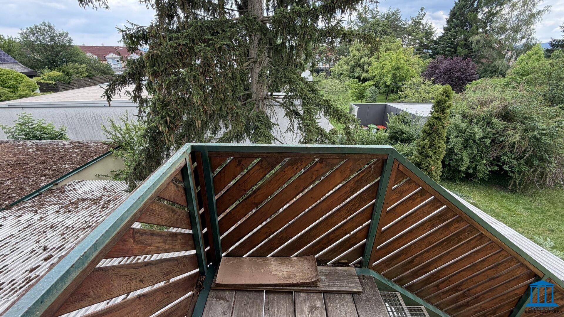 Kleiner Balkon mit Holzboden und Geländer, umgeben von Bäumen und Grünflächen, bietet eine private Außenoase.