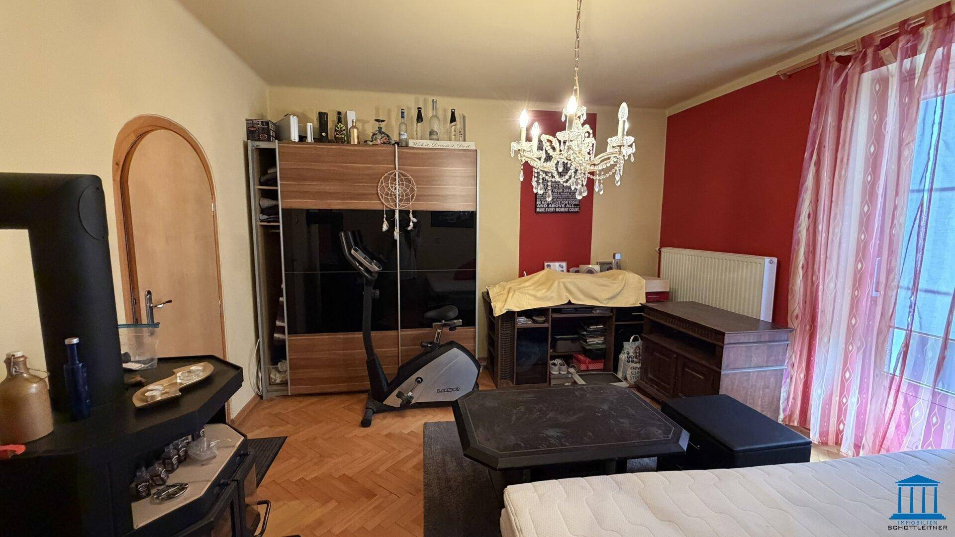 Schlafzimmer mit Doppelbett, Kleiderschrank und Trainingsgerät, roter Akzentwand, bietet vielseitige Nutzungsmöglichkeiten.