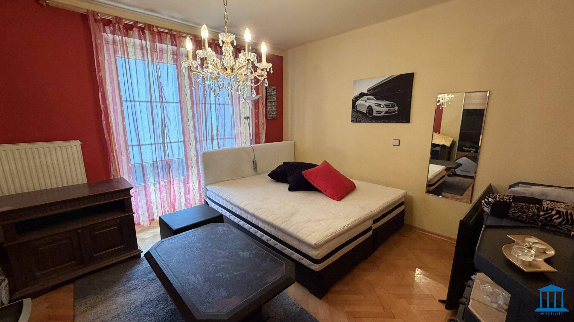 Schlafzimmer mit Doppelbett, roter Akzentwand und Kronleuchter, Fenster mit Vorhängen, schafft eine gemütliche Atmosphäre.