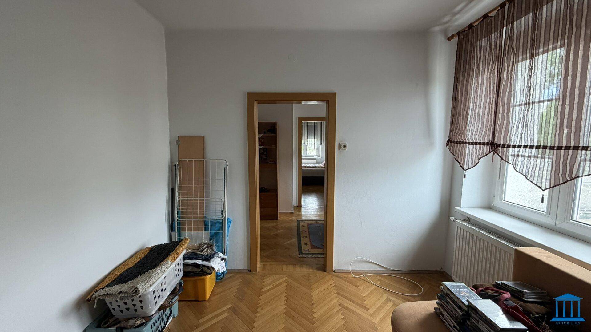 Heller Wohnraum mit Holzboden und Fenster, Blick in einen angrenzenden Raum, vielseitig nutzbar.