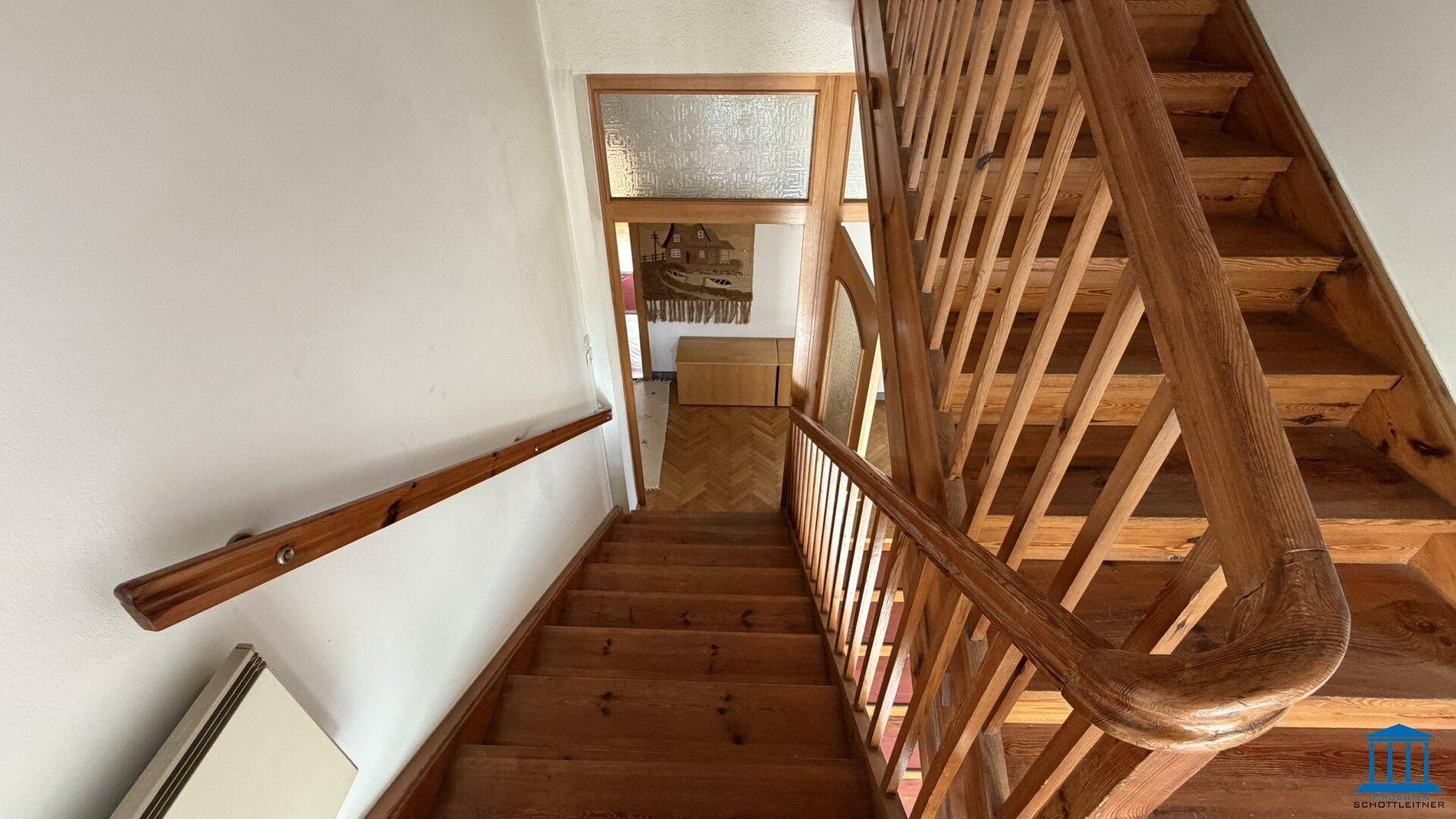 Holztreppe mit Geländer führt in den oberen Bereich, heller Flur im Erdgeschoss, gut erhalten.
