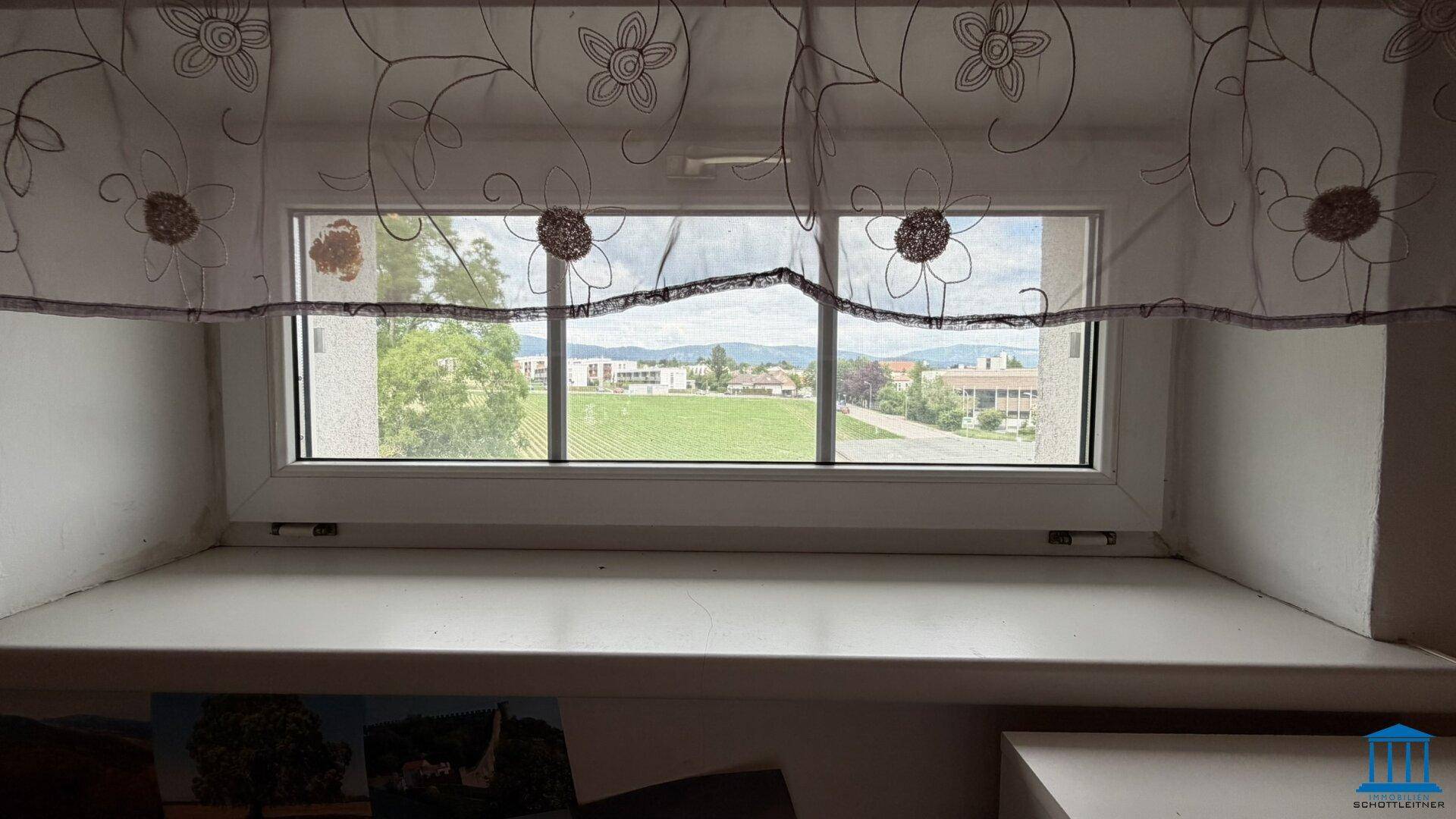 Blick aus einem Fenster mit dekorativen Vorhängen auf eine grüne Landschaft und entfernte Berge.