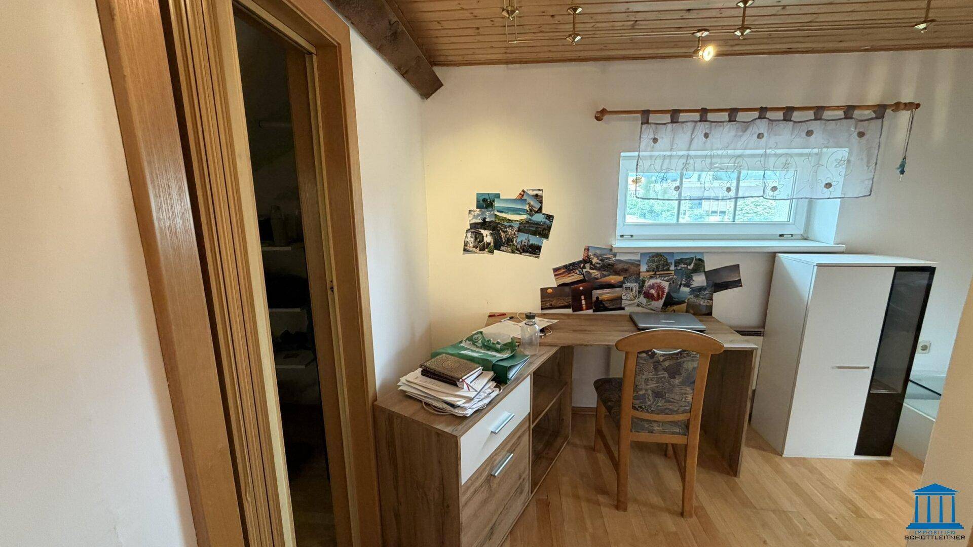 Arbeitsbereich in einem Zimmer mit Holzdecke, Schreibtisch und einem Fenster mit Spitzenvorhang.