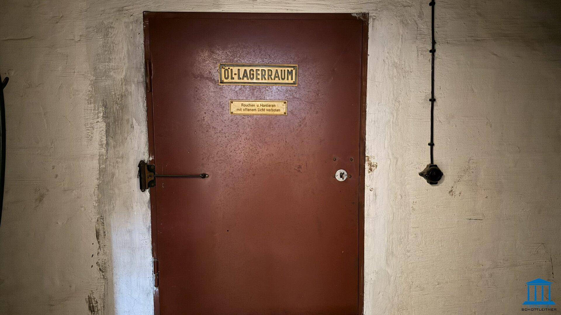 Rote Metalltür in einem Kellerraum, beschriftet als Öl-Lagerraum mit Warnhinweisen.