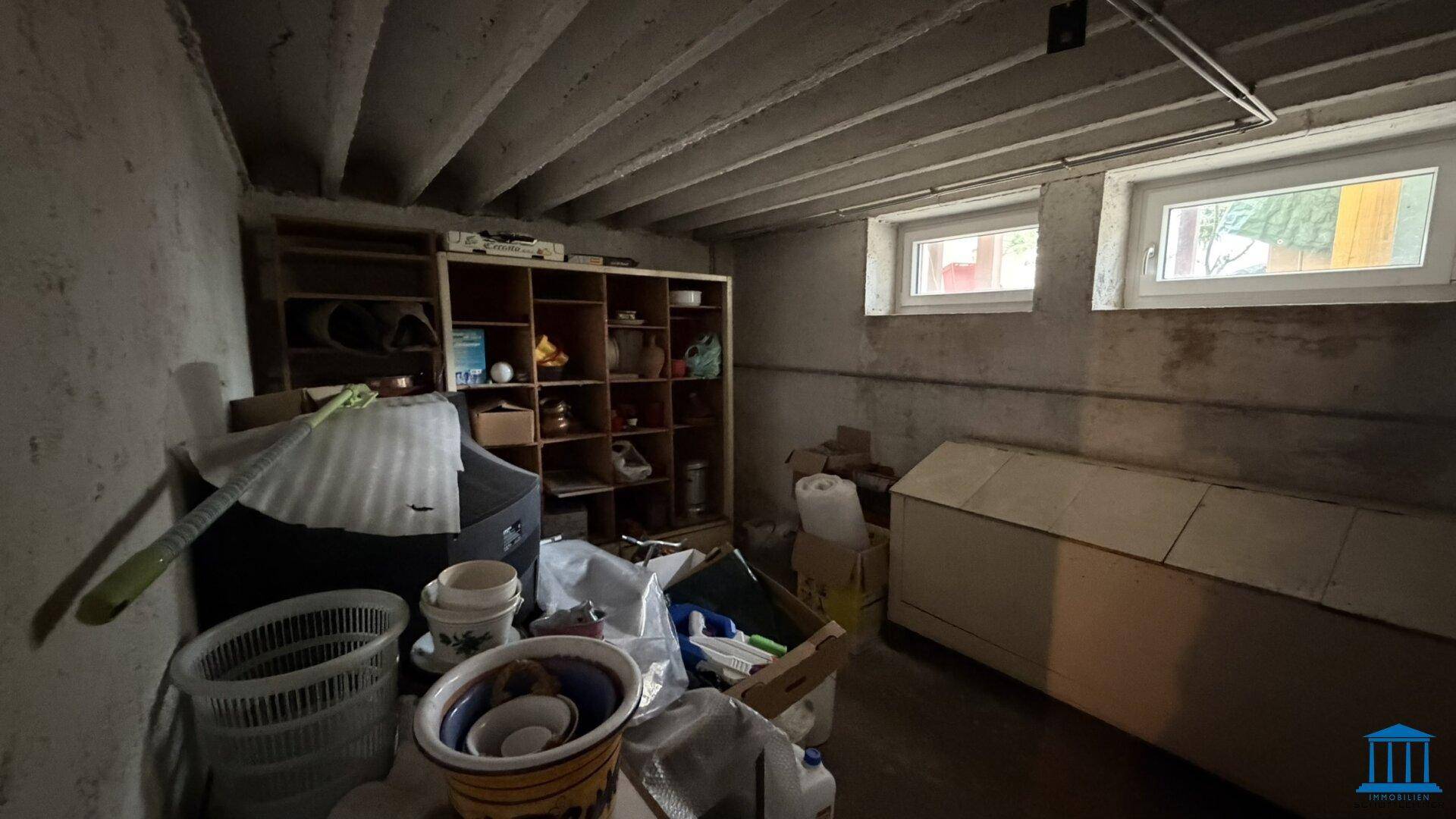 Kellerraum mit Betonbalkendecke, Holzregal und zwei kleinen Fenstern, gefüllt mit verschiedenen Gegenständen.