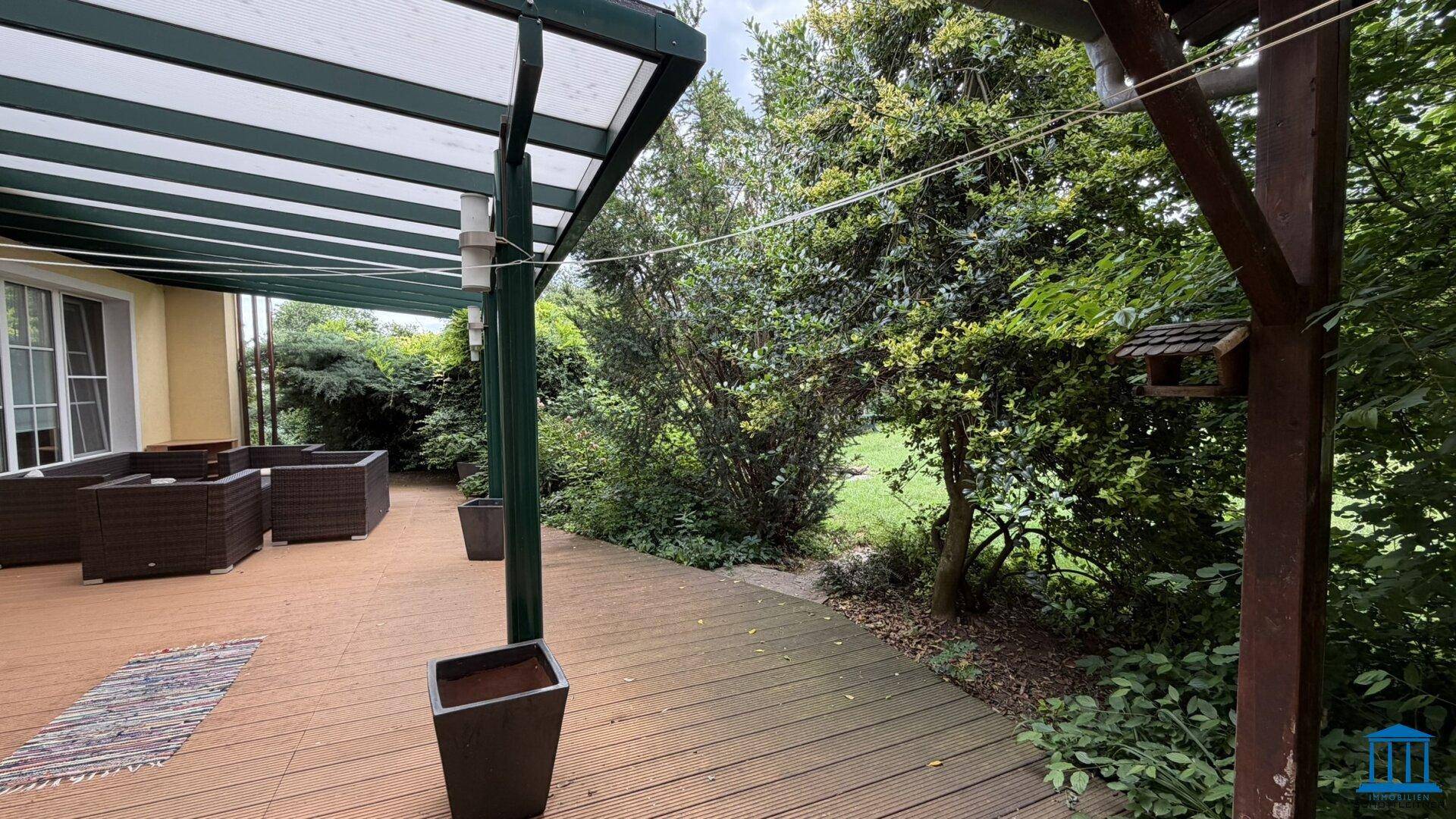 Teil der überdachten Holzterrasse mit angrenzendem Gartenbereich und üppiger Bepflanzung.