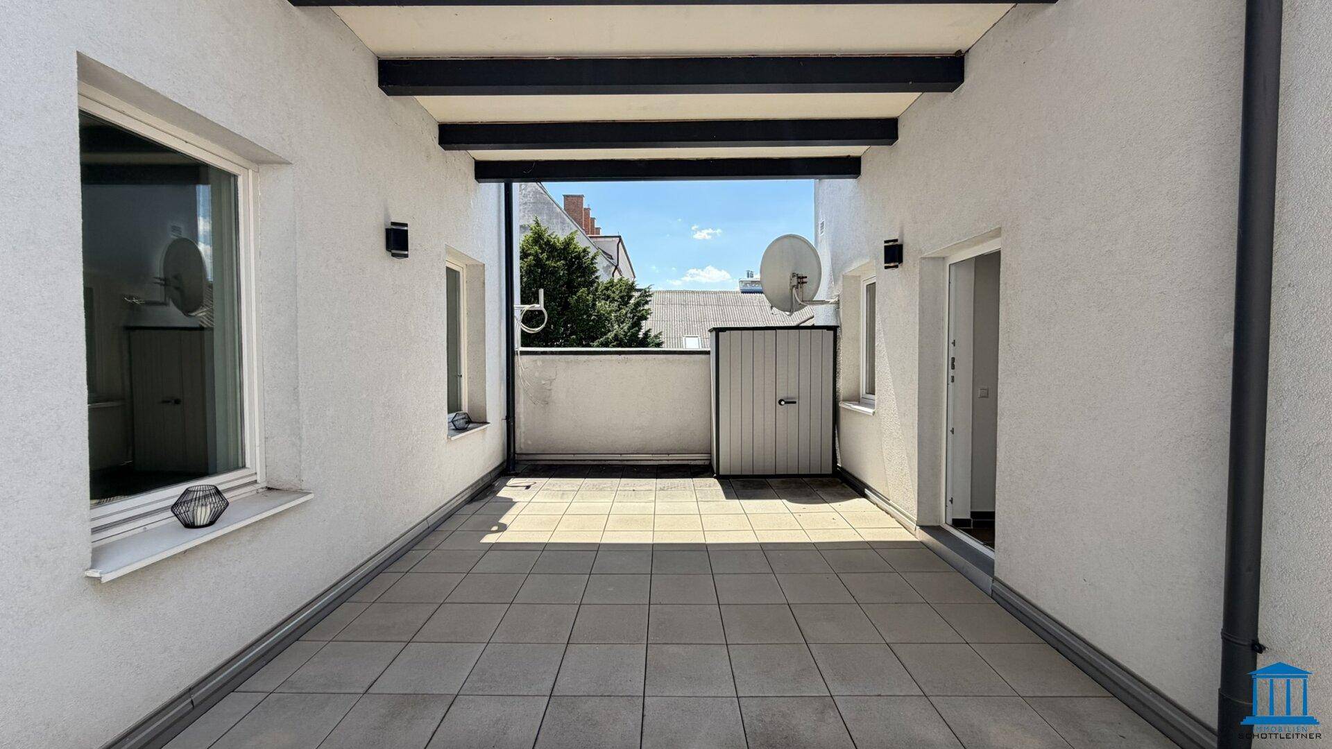Große, geflieste Terrasse mit Überdachung und Blick auf umliegende Gebäude unter blauem Himmel.