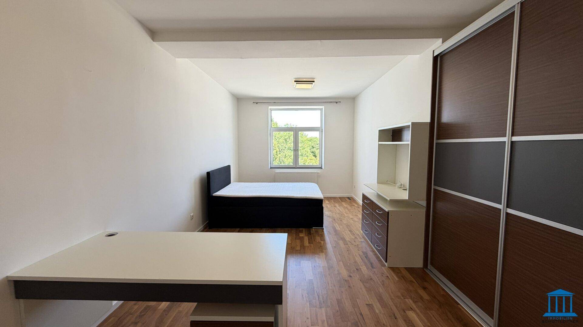 Schlafzimmer mit Holzboden, großem Fenster und einem Bett, sowie einem Schreibtisch.