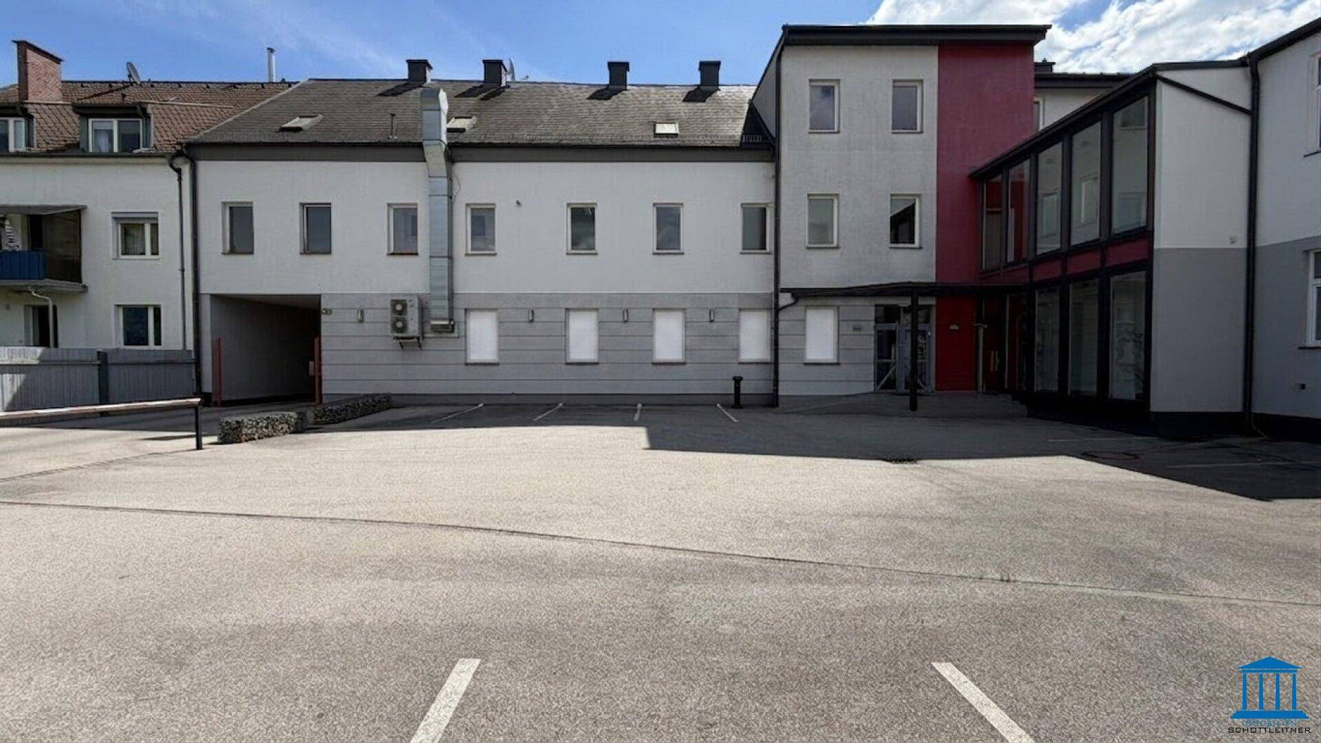 Ansicht der Gebäuderückseite mit Parkplätzen und einem modernen Anbau mit Glasfassade.