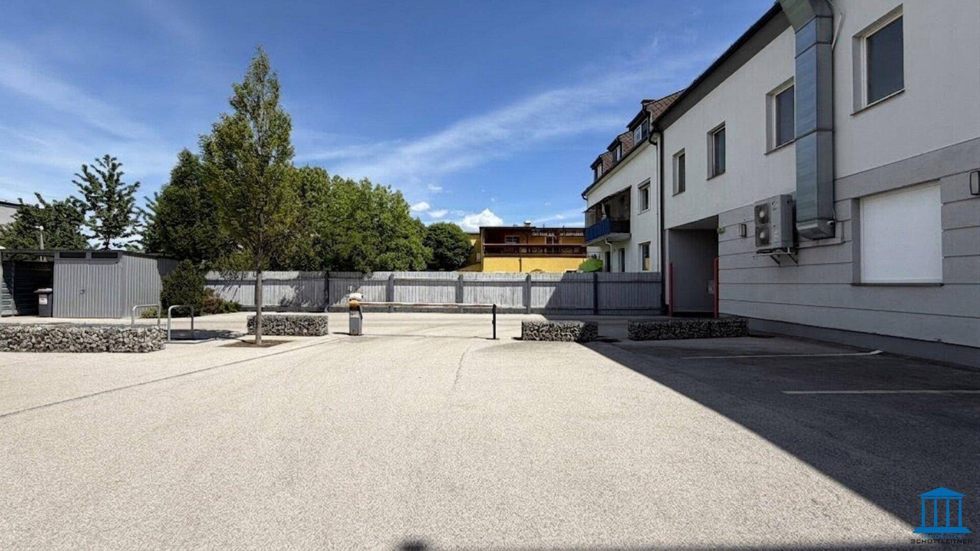 Großer Außenparkplatz mit Bäumen und einem Zaun im Hintergrund, an einem sonnigen Tag.