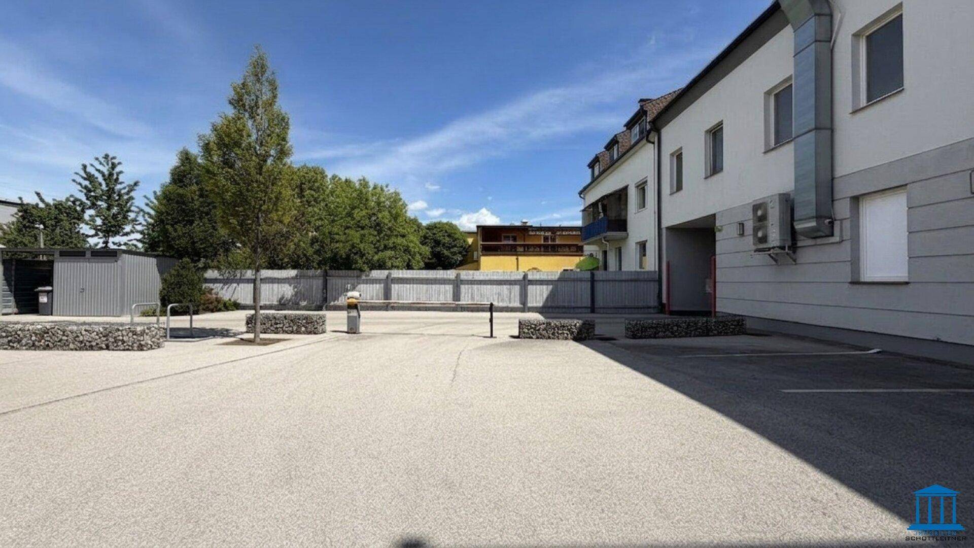 Großer Außenparkplatz mit Bäumen und einer seitlichen Ansicht des Gebäudes unter blauem Himmel.