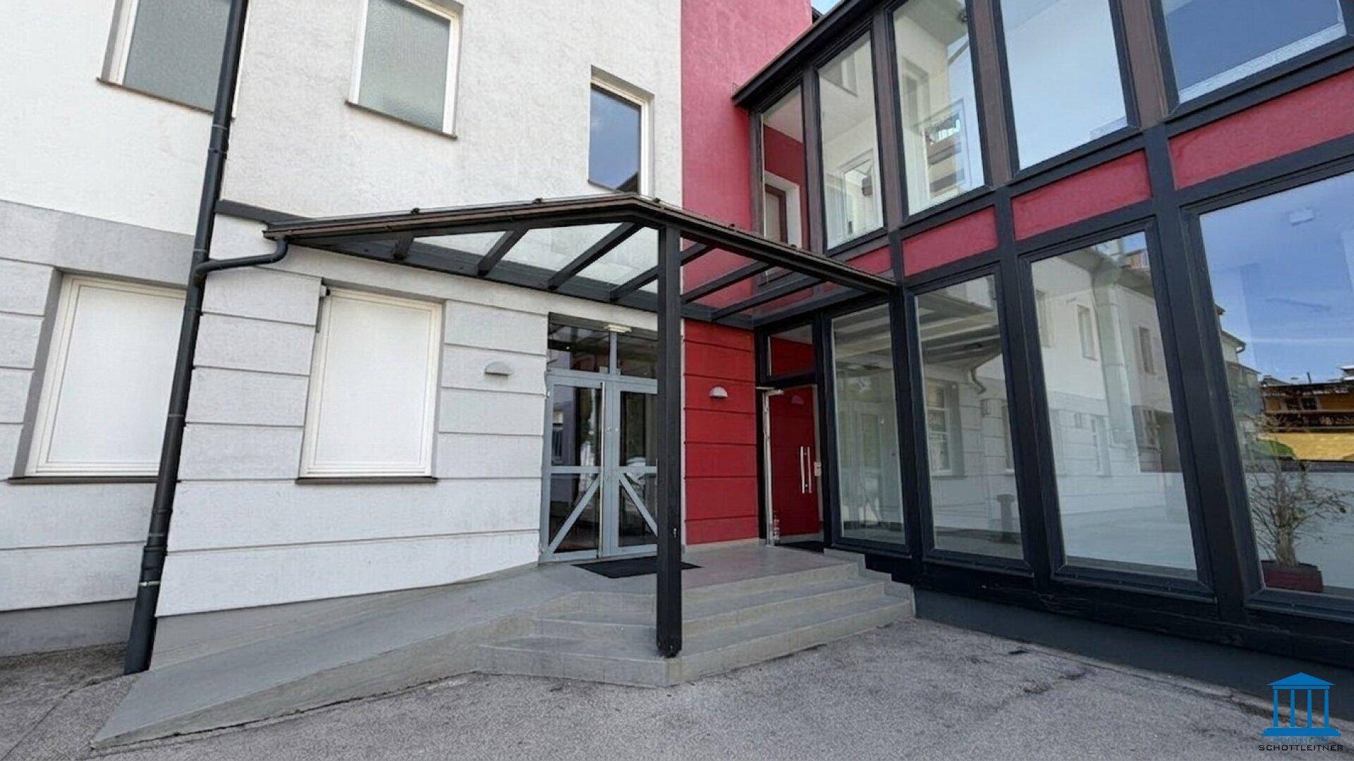 Eingangsbereich eines modernen Gebäudes mit Glasvordach, Stufen und einer barrierefreien Rampe.