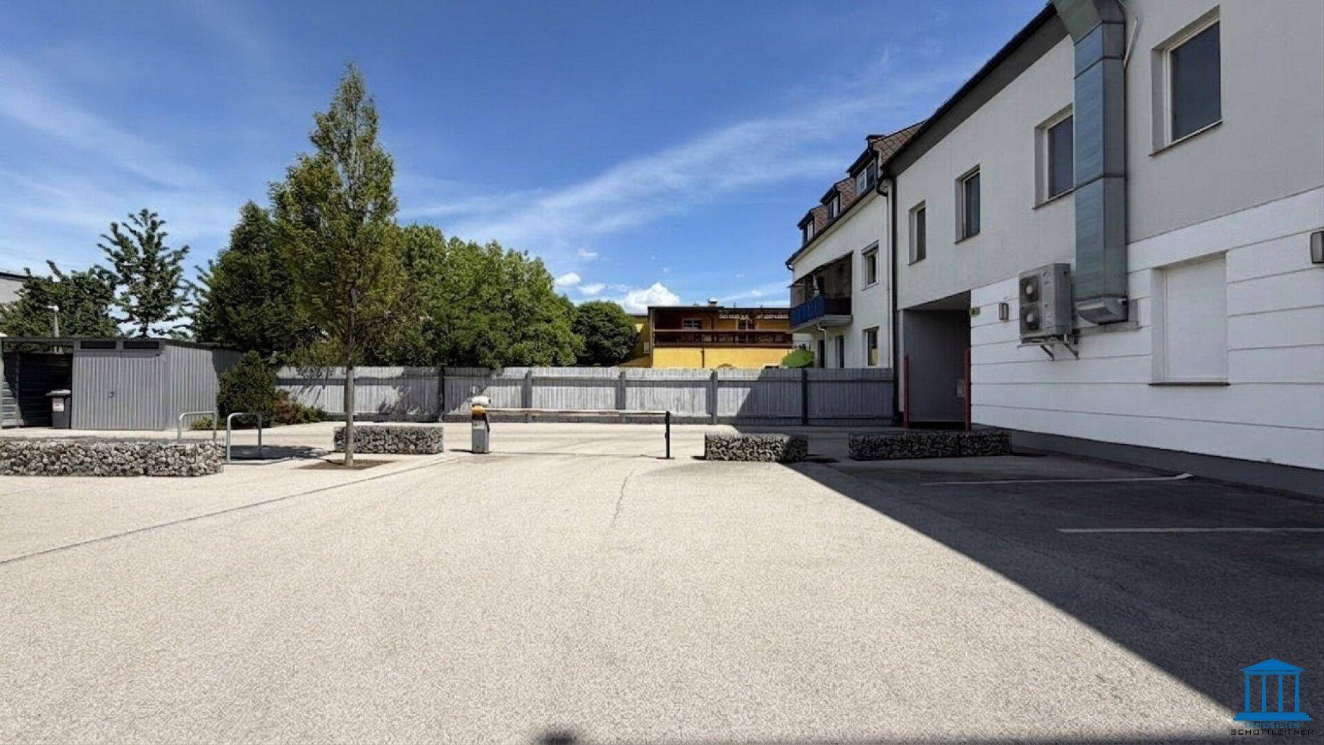 Blick auf den Parkplatz und die Gebäudeseite, umgeben von Bäumen und einem Zaun im Hintergrund.