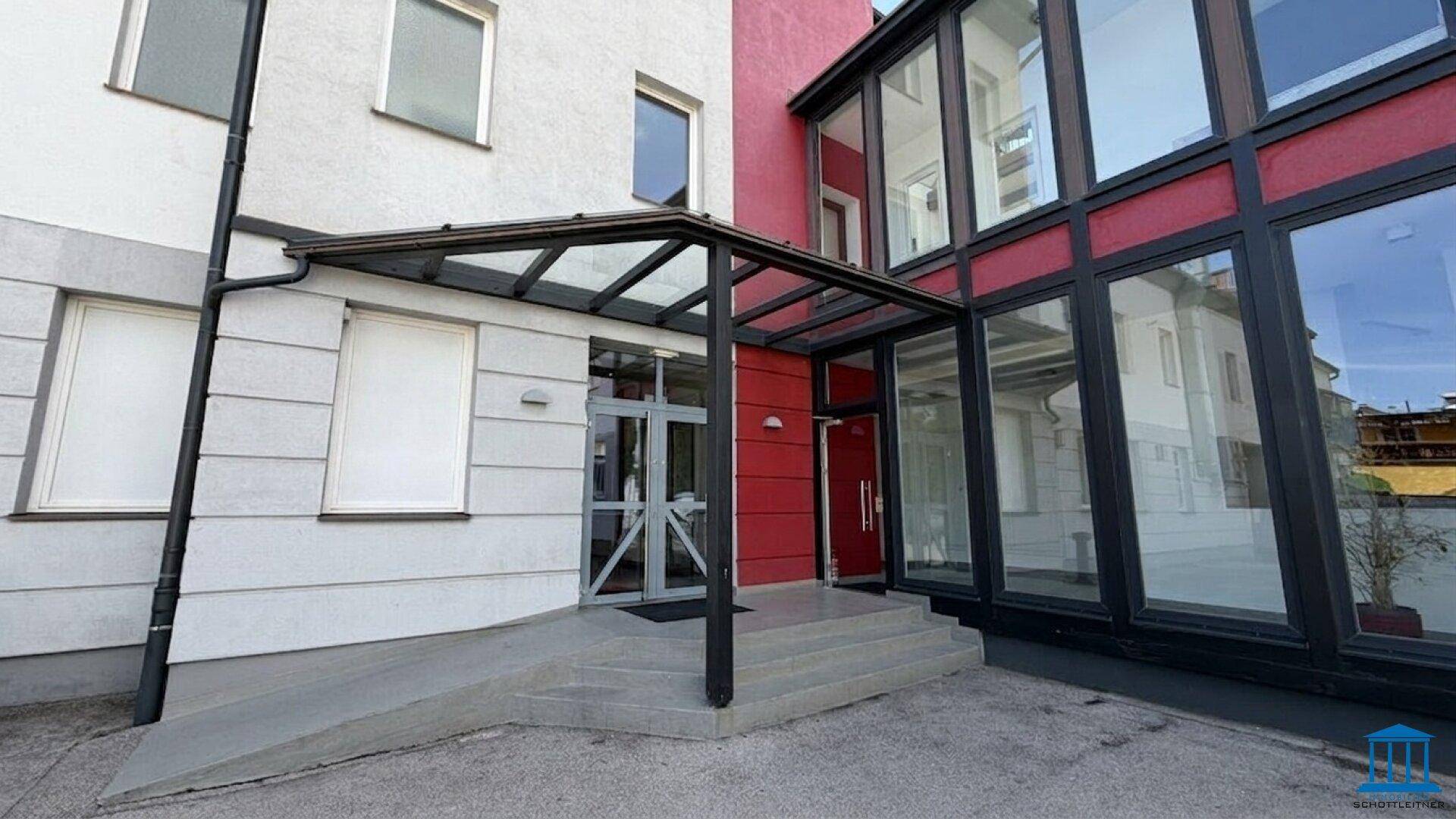 Eingangsbereich eines modernen Gebäudes mit Glasvordach und barrierefreier Rampe, umgeben von weißer und roter Fassade.