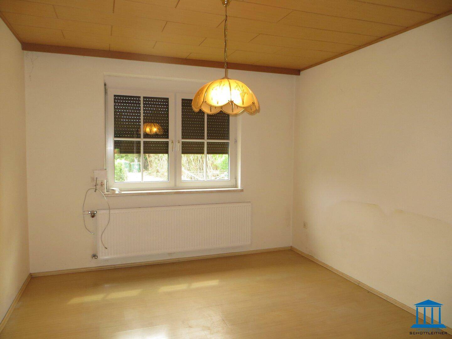 Leeres Zimmer mit Parkettboden, Fenster mit Rollläden, Heizkörper und einer klassischen Deckenleuchte.