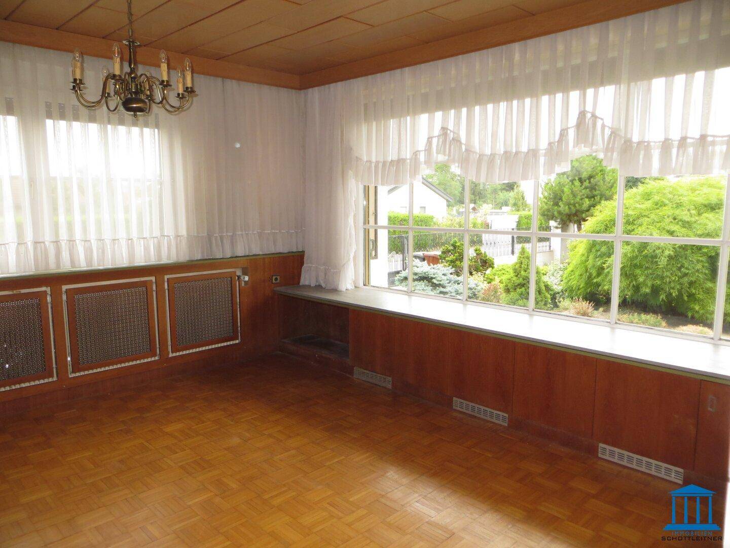 Wohnzimmer mit großen Fenstern, Parkettboden, Holzvertäfelung und einem Kronleuchter, hell und geräumig.
