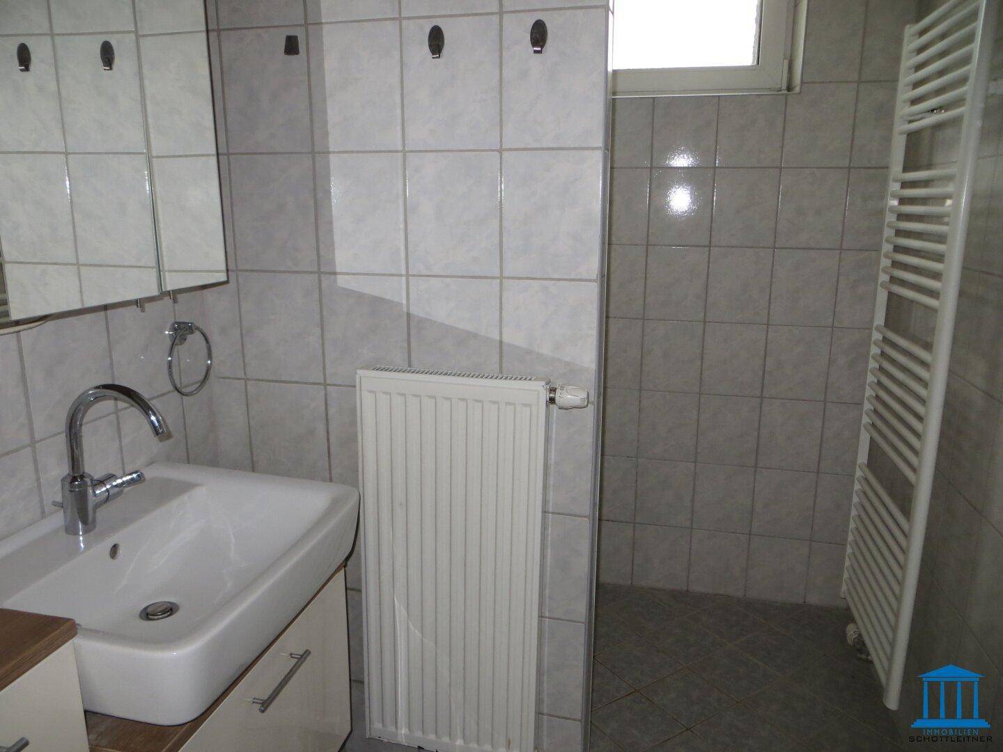 Badezimmer mit grauen Fliesen, Einzelwaschbecken, Duschbereich, Heizkörper und beheiztem Handtuchhalter.