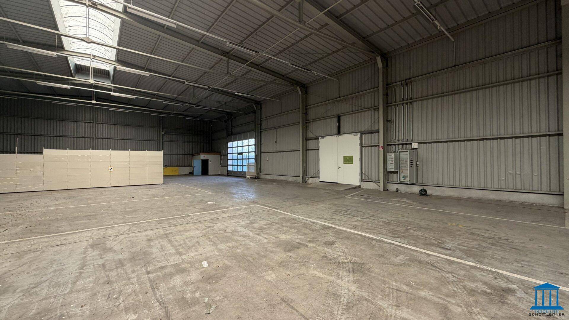 Innenansicht der Lagerhalle mit Betonboden, hoher Decke und einer Tür im hinteren Bereich.