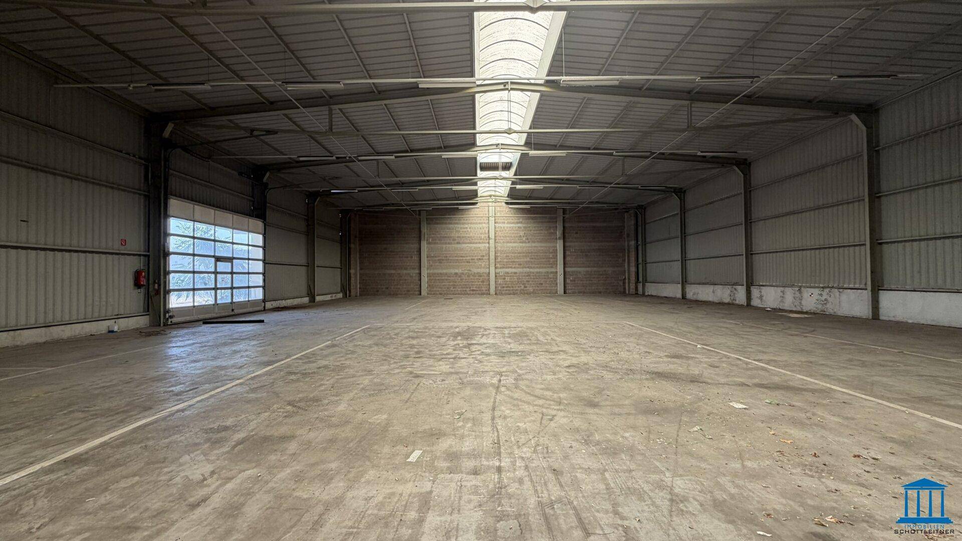 Geräumige, leere Lagerhalle mit hoher Decke, Oberlichtern und einem großen Rolltor.