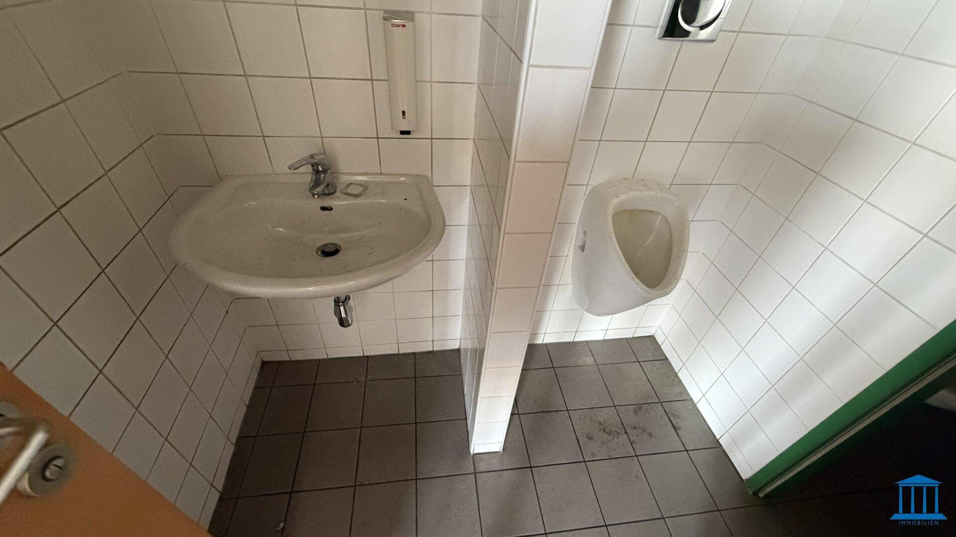 Herren-WC mit Urinal, Waschbecken und weißen Fliesen an Wänden und Boden.