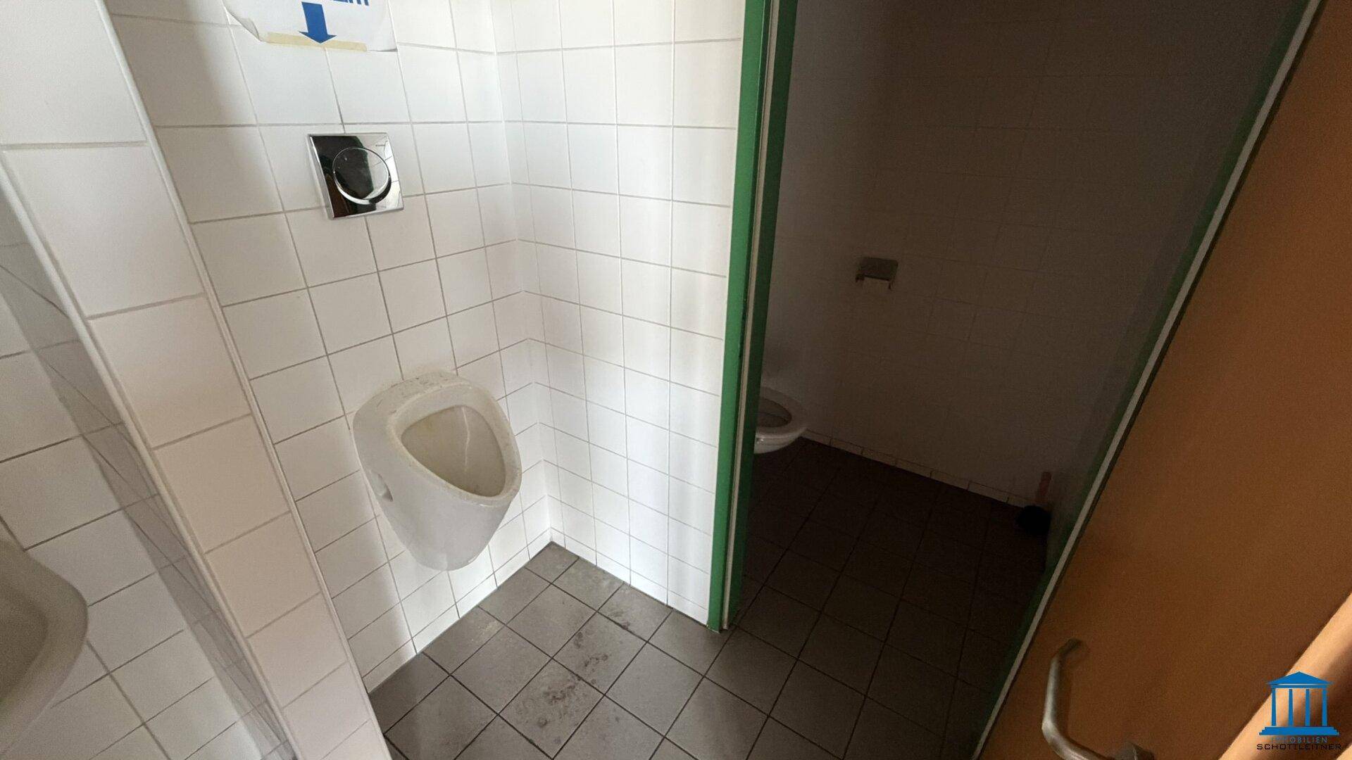 Herren-WC mit Urinal und Toilettenkabine, komplett gefliest in Weiß und Grau.