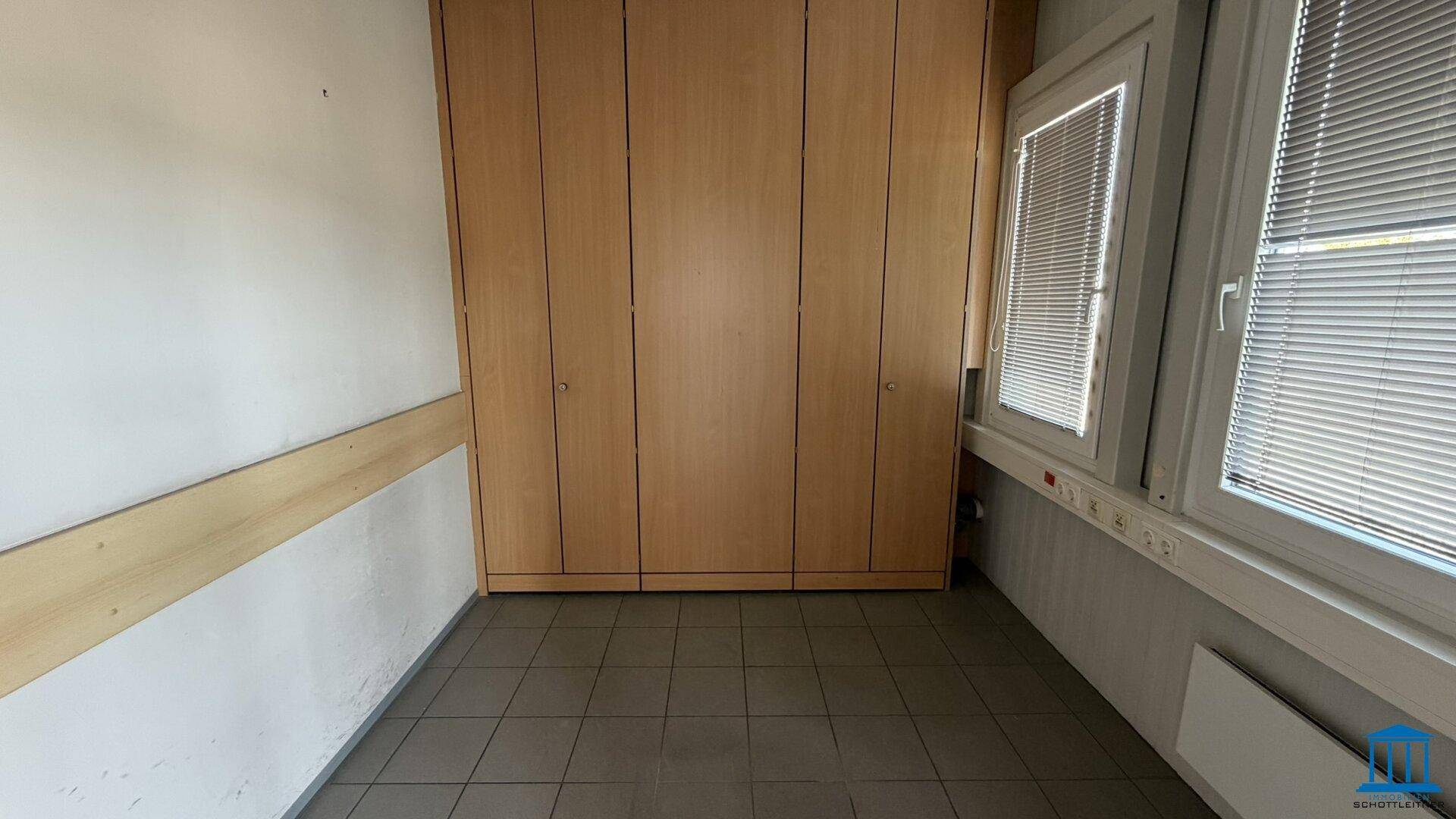 Kleiner Büroraum mit Einbauschränken aus Holz, Fliesenboden und einem Fenster mit Jalousien.