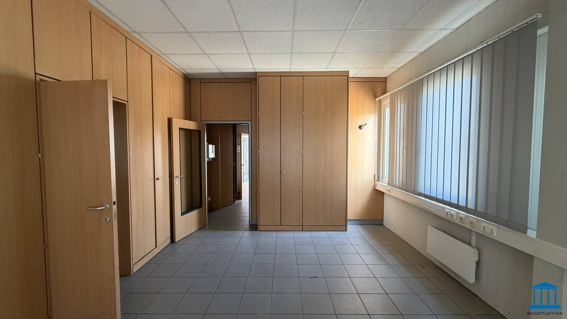 Büroraum mit Einbauschränken aus Holz, Fliesenboden und einem Fenster mit Jalousien für Privatsphäre.