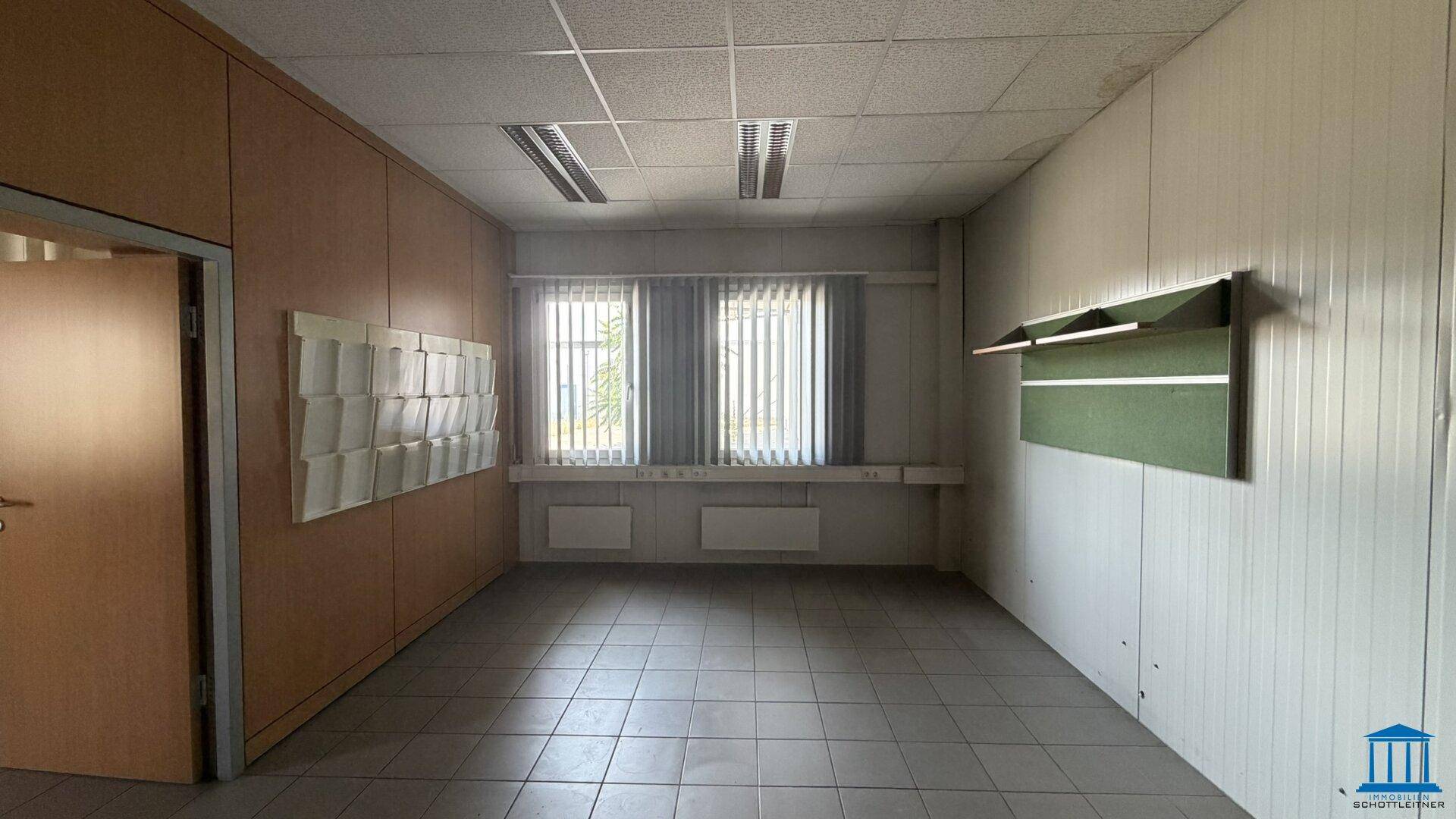 Büroraum mit hellen Wänden, Fliesenboden und zwei Fenstern mit Jalousien, die für ausreichend Licht sorgen.
