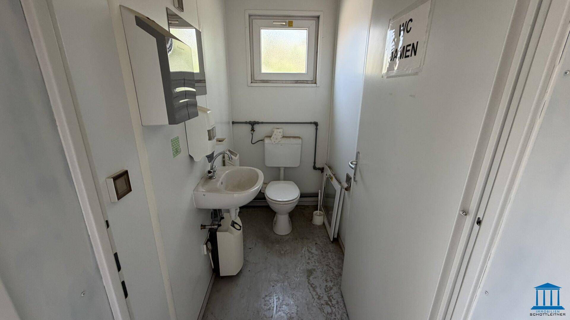 Kleine Toilette in einem Bürocontainer mit WC, Waschbecken und einem Fenster für Belüftung.