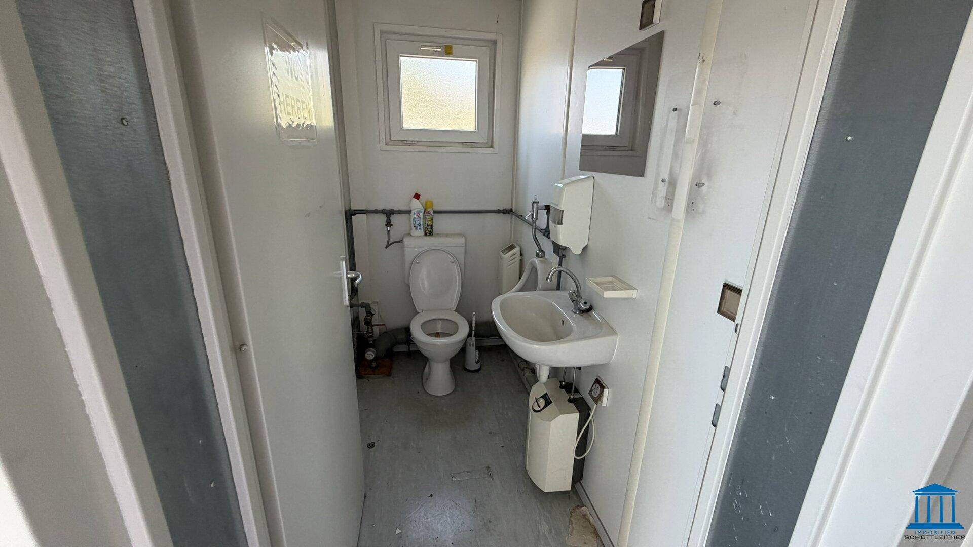Ansicht einer einfachen Toilette in einem Bürocontainer mit WC, Waschbecken und Fenster.