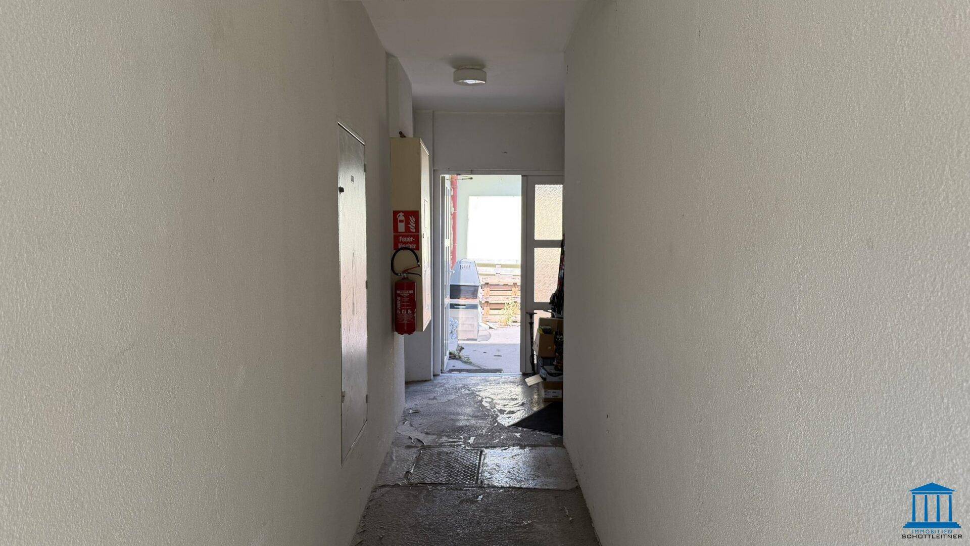 Schmaler Flur mit weißen Wänden, einem Feuerlöscher und Blick auf den Ausgangsbereich.