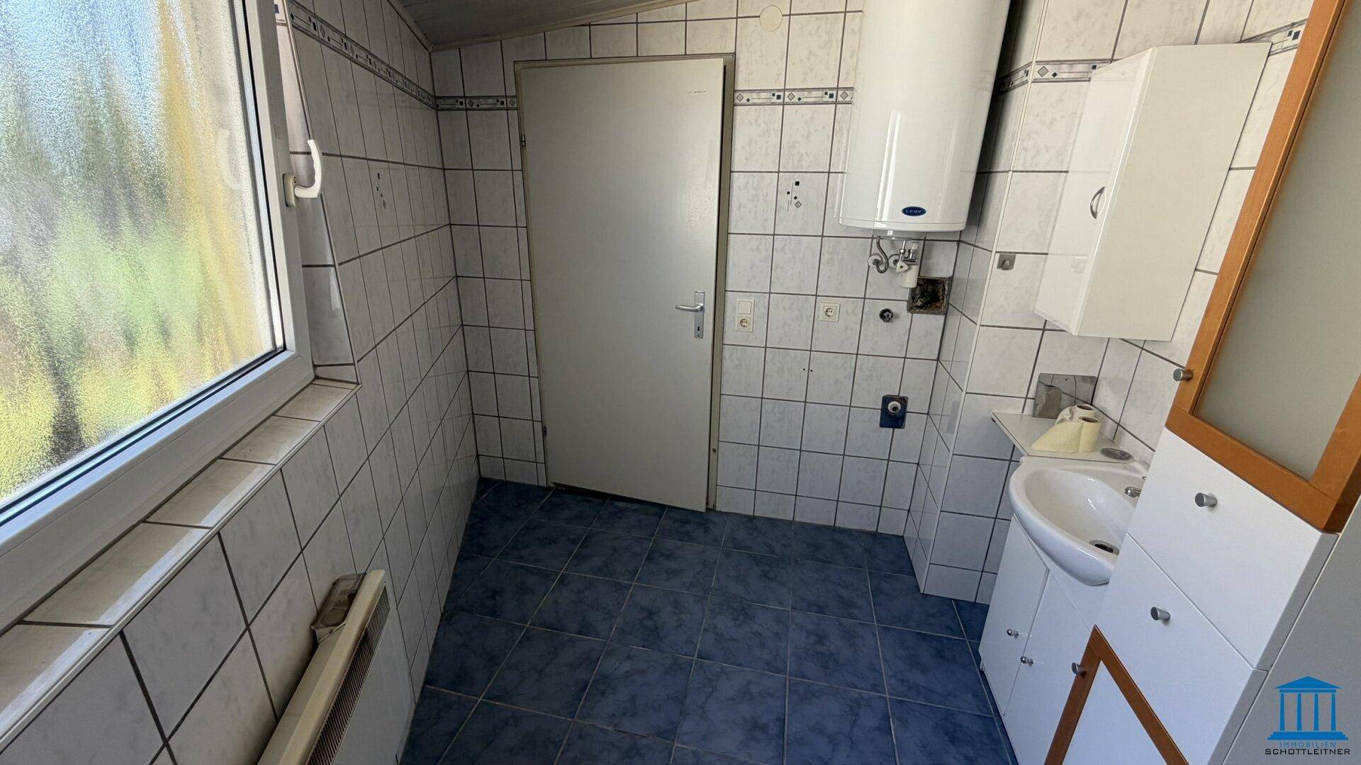 Gefliestes Badezimmer mit Fenster, Waschbecken und einem Warmwasserboiler an der Wand.