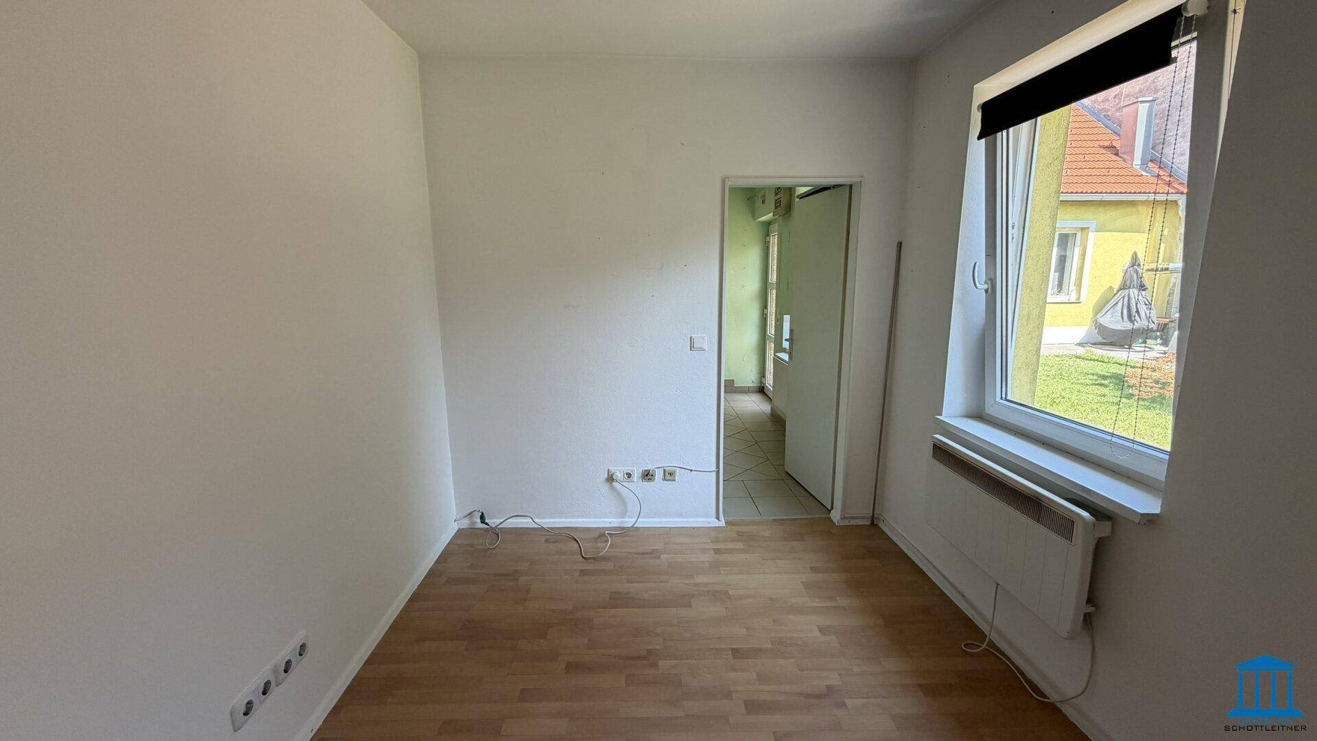 Leeres Zimmer mit Laminatboden, weißen Wänden und einem Fenster mit Blick nach draußen.