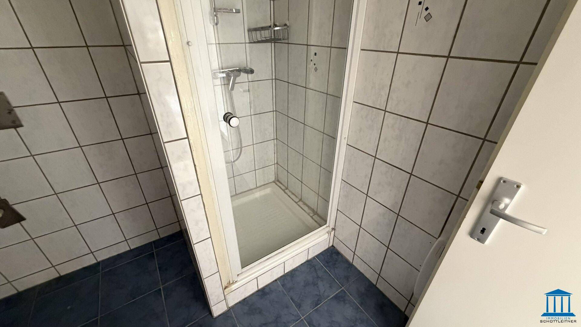Badezimmer mit Duschkabine, weißen Wandfliesen und blauen Bodenfliesen im älteren Stil.