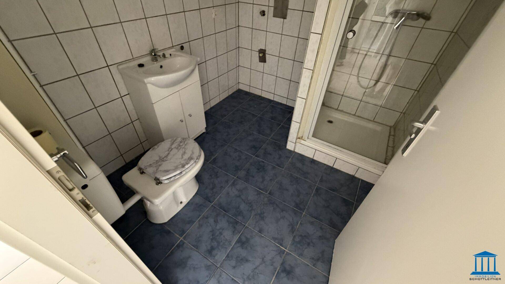 Kleines Badezimmer mit blauen Bodenfliesen, weiß gefliesten Wänden, Toilette und Duschkabine.