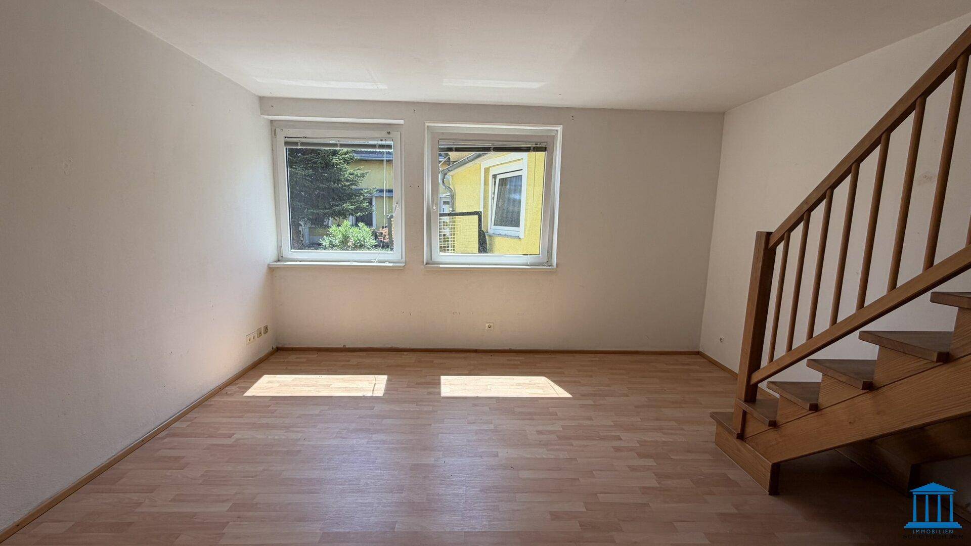 Heller Wohnbereich mit Laminatboden, zwei Fenstern und einer Holztreppe zum Obergeschoss.