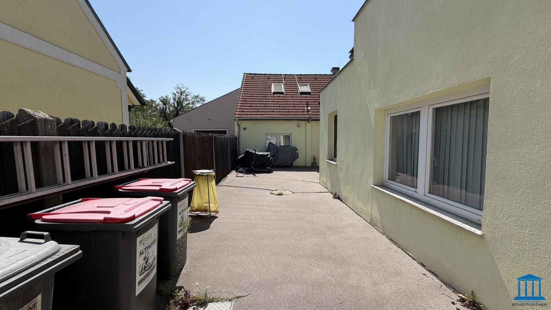 Gepflasterter Zugangsbereich zwischen den Gebäuden mit Mülltonnen und Blick auf das hintere Haus.