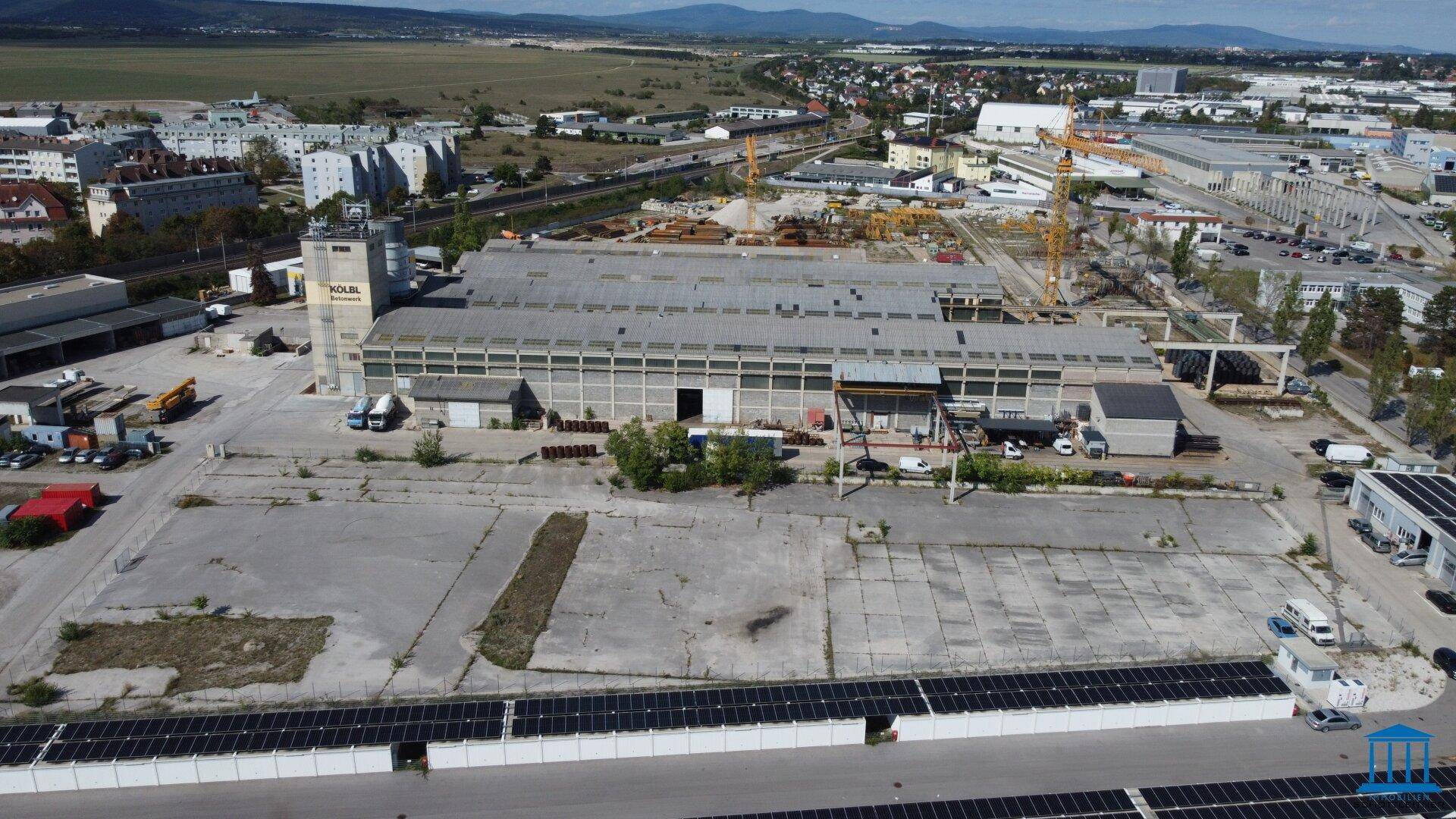 Panoramablick aus der Luft auf ein großes Industriegebäude, Freiflächen, Solaranlagen und die umliegende Landschaft.